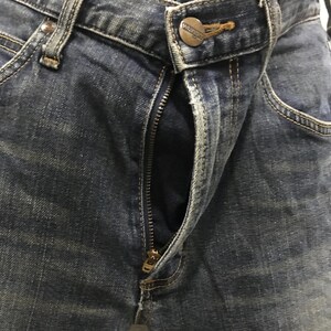 Pu&ograve; includere: Primo piano di jeans blu in denim con cerniera parzialmente aperta. I jeans hanno un classico design a cinque tasche, con cuciture visibili e chiusura con bottone in metallo. Il logo del marchio Wrangler &egrave; visibile.
