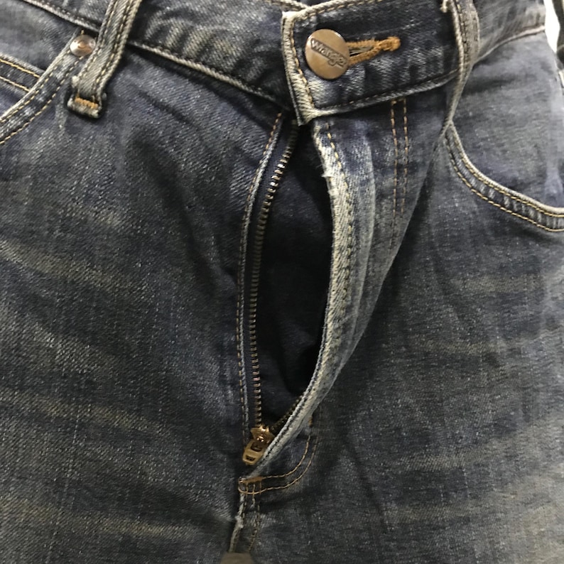 Pu&ograve; includere: Primo piano di jeans blu in denim con cerniera parzialmente aperta. I jeans hanno un classico design a cinque tasche, con cuciture visibili e chiusura con bottone in metallo. Il logo del marchio Wrangler &egrave; visibile.