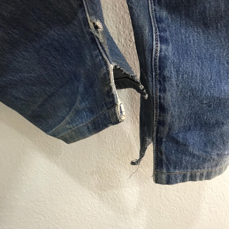 May include: Close-up of blue denim jeans with distressed details. The jeans show signs of wear and tear, with frayed edges and a ripped seam. The denim has a faded appearance, suggesting it has been washed multiple times.