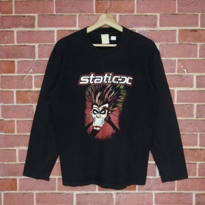 Puede incluir: Camiseta negra de manga larga con un estampado gráfico de un personaje de dibujos animados con pelo rojo y amarillo, una boca roja y un fondo rojo y negro. El texto "staticx" está impreso en blanco sobre el personaje.