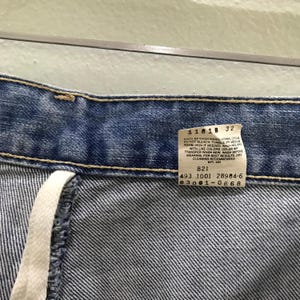 May include: Blue denim jeans featuring a white interior care label with black text that reads 1181B 32, 821, 493 1001 28984-6, and 03001-0668. The jeans have a visible waistband with yellow stitching and a white fabric drawstring hanging from the side.