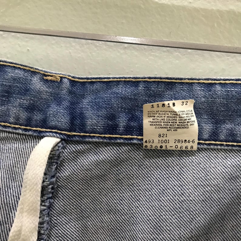 May include: Blue denim jeans featuring a white interior care label with black text that reads 1181B 32, 821, 493 1001 28984-6, and 03001-0668. The jeans have a visible waistband with yellow stitching and a white fabric drawstring hanging from the side.