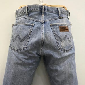 May include: Light blue denim jeans with the Wrangler logo on a brown leather patch. The jeans have classic five-pocket styling and a straight leg fit. The back pockets feature a signature W-shaped stitching.