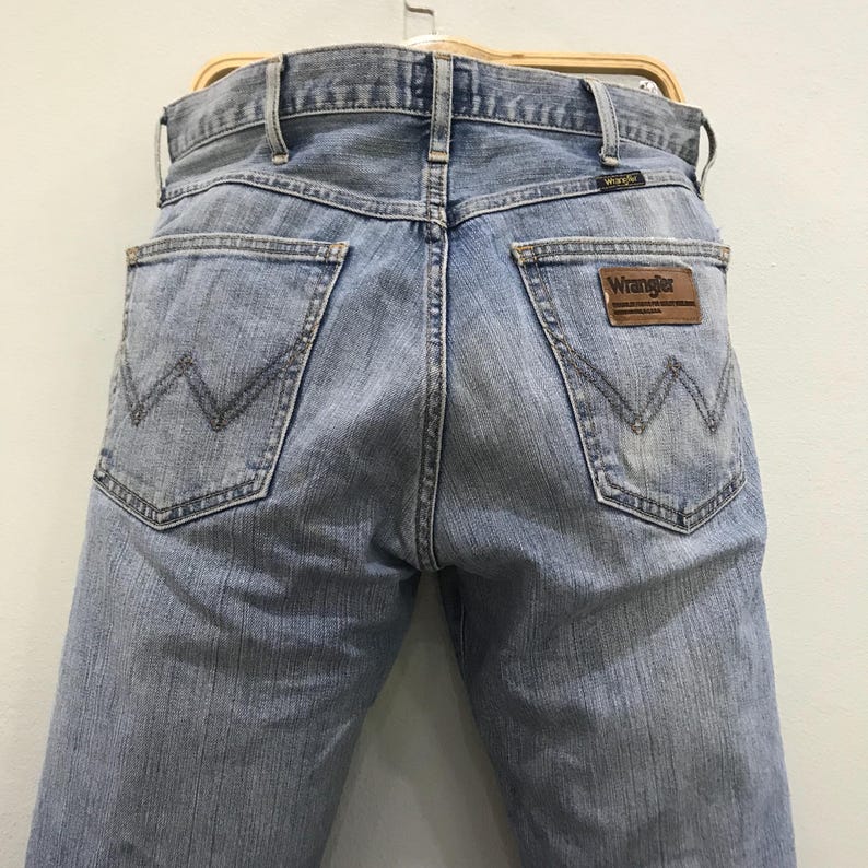 May include: Light blue denim jeans with the Wrangler logo on a brown leather patch. The jeans have classic five-pocket styling and a straight leg fit. The back pockets feature a signature W-shaped stitching.