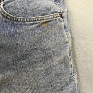 May include: Close-up of light blue denim jeans. The jeans have a pocket with a brown stain and a small brown stain on the leg. The denim has a faded appearance.