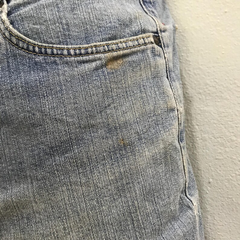 May include: Close-up of light blue denim jeans. The jeans have a pocket with a brown stain and a small brown stain on the leg. The denim has a faded appearance.
