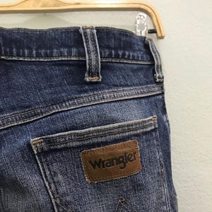 May include: Close-up of blue denim jeans with a brown leather Wrangler patch on the back pocket. The jeans are hanging on a wooden hanger. The denim has a faded, worn appearance.