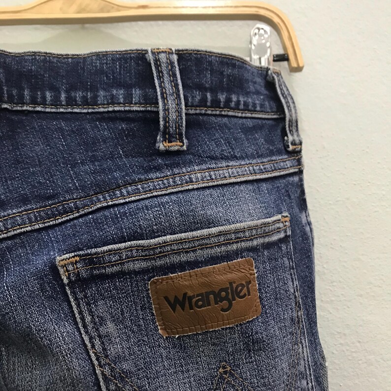 May include: Close-up of blue denim jeans with a brown leather Wrangler patch on the back pocket. The jeans are hanging on a wooden hanger. The denim has a faded, worn appearance.
