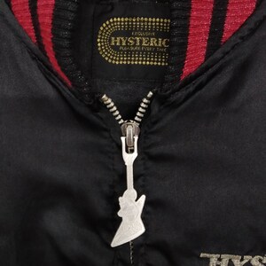 Może przedstawiać: Czarna satynowa kurtka bomberowa z czerwono-czarną paskowaną st&oacute;jką. Suwak ma kształt gitary. Etykieta na kurtce głosi "Exclusive Hysteric Pleasure Every Time".