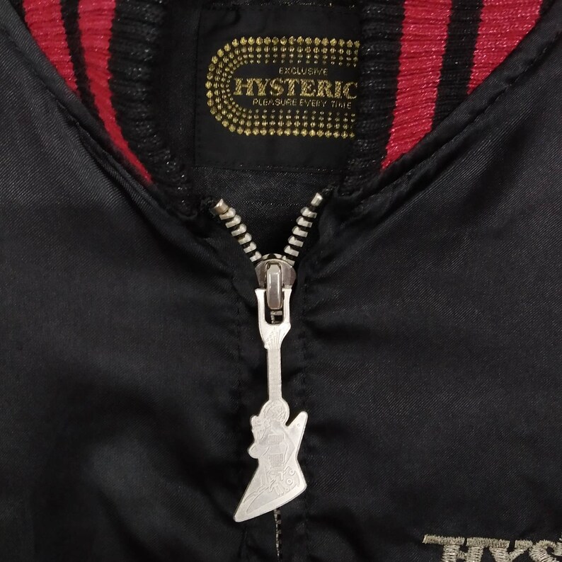 Może przedstawiać: Czarna satynowa kurtka bomberowa z czerwono-czarną paskowaną st&oacute;jką. Suwak ma kształt gitary. Etykieta na kurtce głosi "Exclusive Hysteric Pleasure Every Time".