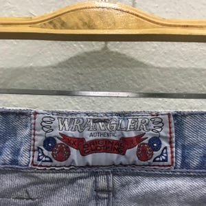 May include: Close-up of light blue denim jeans with a "Wrangler" label. The label is white with red and blue accents, and the words "Authentic Original Clothing" are visible.