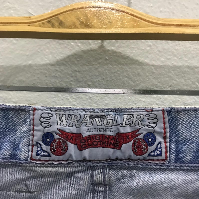 May include: Close-up of light blue denim jeans with a "Wrangler" label. The label is white with red and blue accents, and the words "Authentic Original Clothing" are visible.