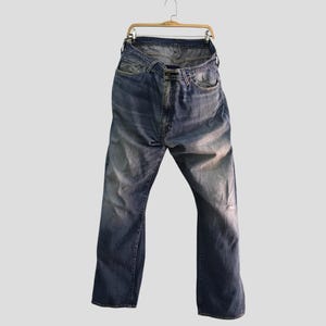 May include: Faded blue denim jeans hanging on a wooden hanger. The jeans have a classic five-pocket design, a button and zipper closure, and a distressed look with a small tear on the right leg. The jeans are a medium wash.