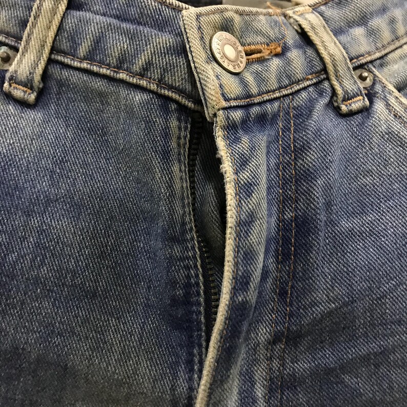 以下が含まれることがあります： ボタン留めとジッパーが付いた、青いデニムジーンズのクローズアップ。ジーンズは色あせたウォッシュ加工で、ステッチのディテールが見えます。ボタンは円形のデザインのシルバーです。