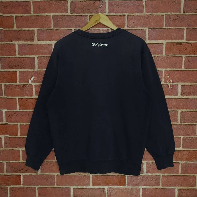 May include: Black sweatshirt with long sleeves and a crew neck. The back of the sweatshirt has the text "&copy; K. Haring" printed in white.