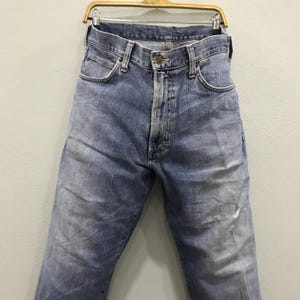 May include: Light blue denim jeans hanging on a wooden hanger. The jeans have a classic five-pocket design, a button and zipper closure, and show signs of wear with faded areas and a small tear near the knee.