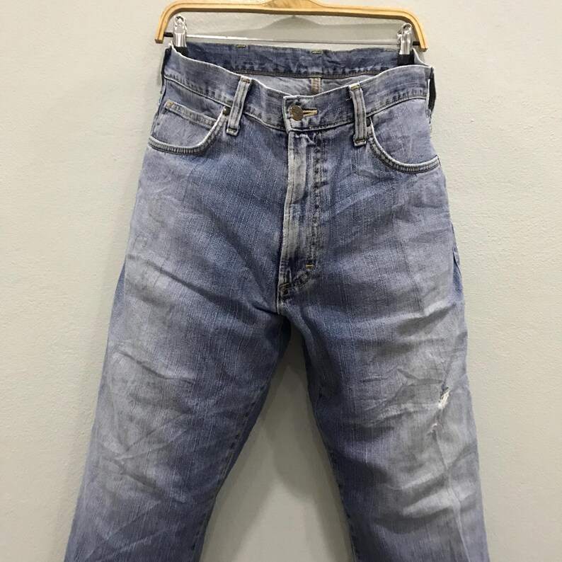 May include: Light blue denim jeans hanging on a wooden hanger. The jeans have a classic five-pocket design, a button and zipper closure, and show signs of wear with faded areas and a small tear near the knee.