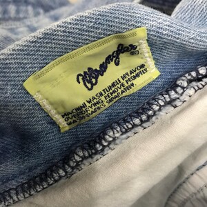 May include: Close-up of a light blue denim fabric with a yellow label. The label features the brand name "Wrangler" in a cursive font and washing instructions. The fabric is detailed with visible stitching.