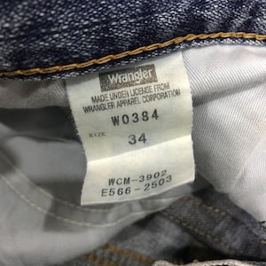 May include: Close-up of a white tag from Wrangler jeans. The tag includes the brand name, size 34, and other details such as "MADE UNDER LICENSE FROM WRANGLER APPAREL CORPORATION", and the codes W0384, WCM-3902, and E566-2503.