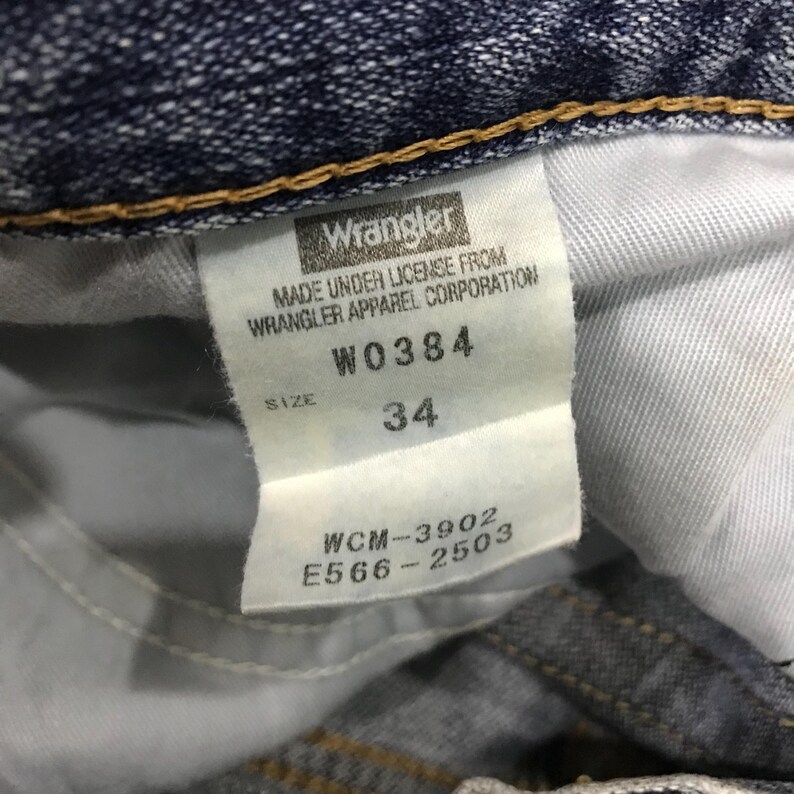 May include: Close-up of a white tag from Wrangler jeans. The tag includes the brand name, size 34, and other details such as "MADE UNDER LICENSE FROM WRANGLER APPAREL CORPORATION", and the codes W0384, WCM-3902, and E566-2503.