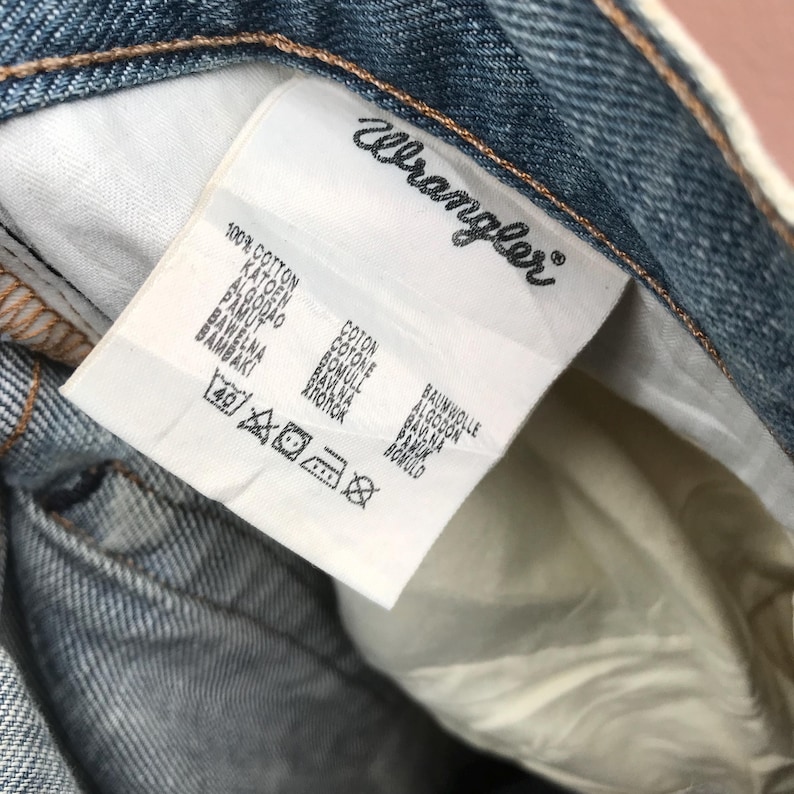 May include: Close-up of a denim garment's interior, showcasing a white tag with the "Wrangler" logo and various fabric care symbols. The tag also lists "100% COTTON" in multiple languages.