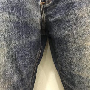 May include: Close-up of blue denim jeans. The jeans have a classic five-pocket design with a button and zip closure. The denim has a faded, worn look, with lighter areas and subtle texture variations.
