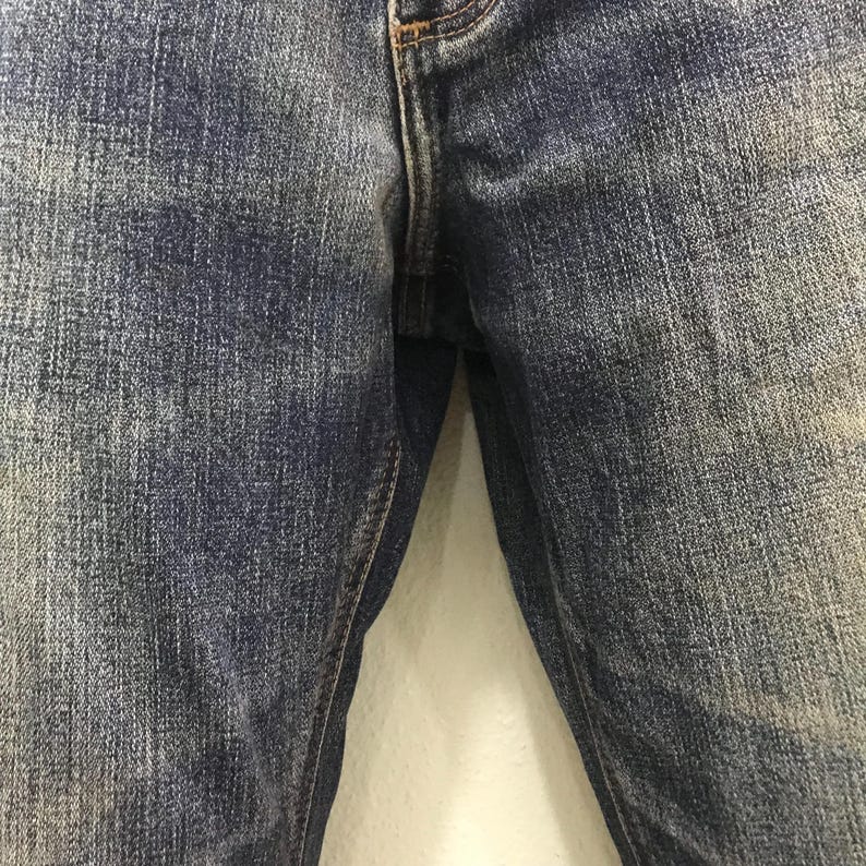 May include: Close-up of blue denim jeans. The jeans have a classic five-pocket design with a button and zip closure. The denim has a faded, worn look, with lighter areas and subtle texture variations.