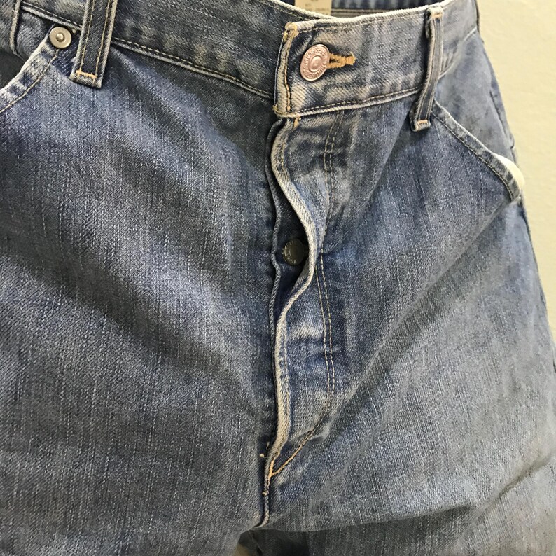 May include: A pair of medium-wash blue denim jeans featuring a classic five-pocket design, a metal button fly closure, and a single metallic button at the waistband. The fabric displays a subtle faded texture with visible stitching along the seams.
