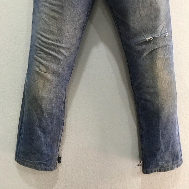 May include: Faded blue denim jeans with a distressed look. The jeans have a worn appearance with light discoloration and a small tear on the right knee. The bottom hems are slightly frayed.