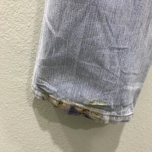 May include: Close-up of the hem of light blue denim jeans. The bottom edge shows wear and discoloration, with a faded appearance. The jeans are slightly wrinkled, indicating they have been worn.