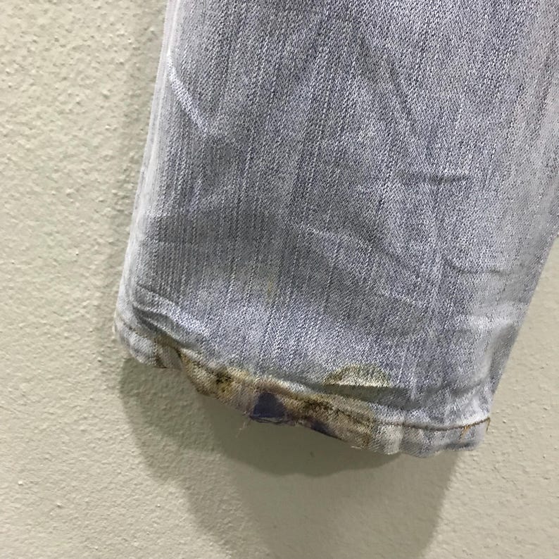May include: Close-up of the hem of light blue denim jeans. The bottom edge shows wear and discoloration, with a faded appearance. The jeans are slightly wrinkled, indicating they have been worn.