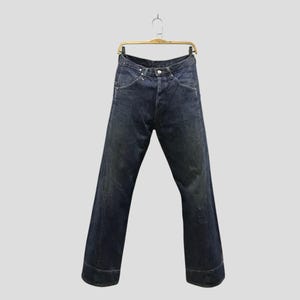 May include: Dark indigo blue denim jeans featuring a high-waisted cut, a button fly closure, and wide legs with prominent stitched cuffs at the bottom. The jeans are displayed hanging from a gold-toned metal hanger against a plain background.