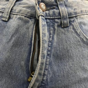 May include: Close-up of light blue denim jeans. The image highlights the zipper, button, and stitching details. The button has a circular design with the letter "M" in the center. The jeans appear to be pre-worn with a faded appearance.