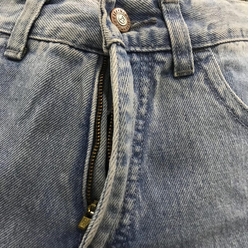 May include: Close-up of light blue denim jeans. The image highlights the zipper, button, and stitching details. The button has a circular design with the letter "M" in the center. The jeans appear to be pre-worn with a faded appearance.