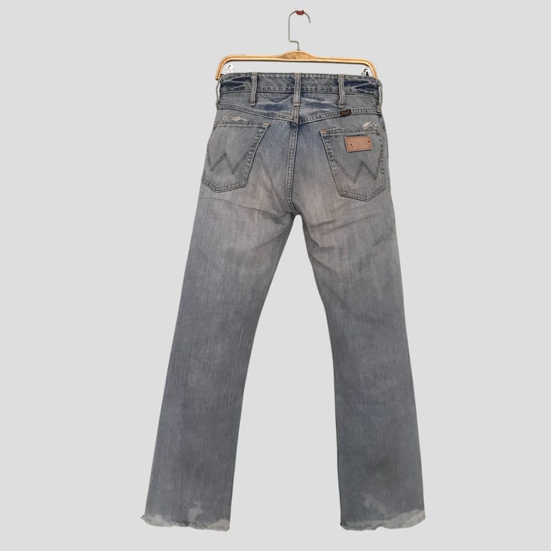 K&ouml;nnte beinhalten: Hellblaue Jeans aus Denim mit verwaschener Optik und Used-Details. Die Jeans h&auml;ngen an einem Holzb&uuml;gel und zeigen die Ges&auml;&szlig;taschen und einen Lederaufn&auml;her. Die Jeans haben ein klassisches F&uuml;nf-Taschen-Design und einen geraden Beinverlauf.