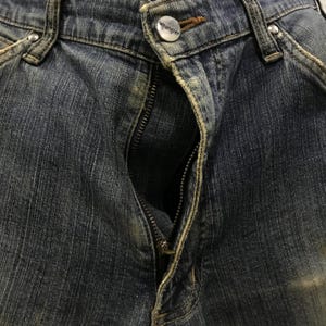 以下が含まれることがあります： 部分的にジッパーが開いた青いデニムジーンズのクローズアップ。ジーンズは、ブランド名「Wrangler」が見えるボタン留めです。デニム生地は、クラシックな5ポケットデザインで、摩耗や色あせの兆候を示しています。
