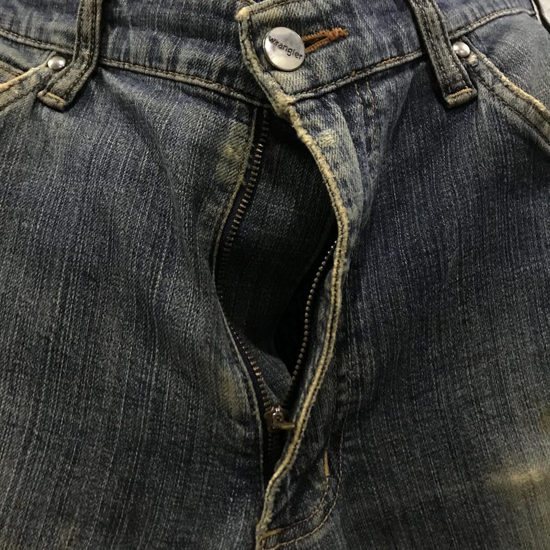 以下が含まれることがあります： 部分的にジッパーが開いた青いデニムジーンズのクローズアップ。ジーンズは、ブランド名「Wrangler」が見えるボタン留めです。デニム生地は、クラシックな5ポケットデザインで、摩耗や色あせの兆候を示しています。