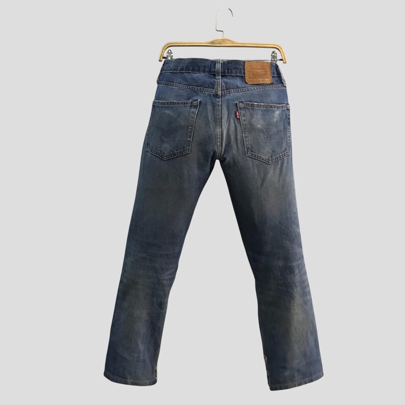 May include: Faded blue denim jeans hanging on a wooden hanger. The jeans have a classic five-pocket design, a brown leather patch, and a red tab on the back pocket. The jeans are slightly distressed.