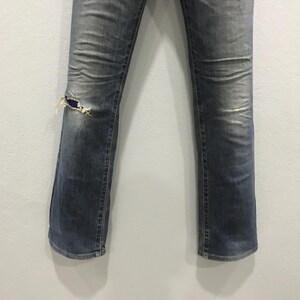 Pu&ograve; includere: Jeans in denim blu sbiadito con ginocchio strappato e usurato. I jeans hanno un classico design a cinque tasche e una gamba leggermente svasata. Il denim mostra segni di usura e scolorimento.