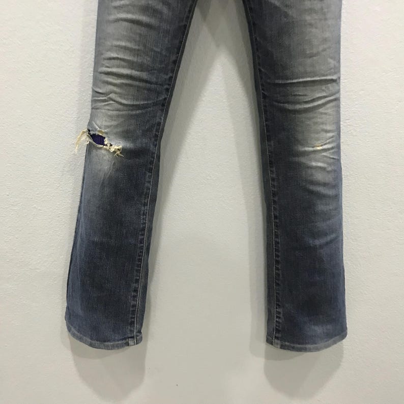 Pu&ograve; includere: Jeans in denim blu sbiadito con ginocchio strappato e usurato. I jeans hanno un classico design a cinque tasche e una gamba leggermente svasata. Il denim mostra segni di usura e scolorimento.