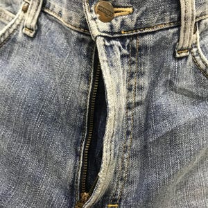 May include: Close-up of blue denim jeans with a visible zipper and a button closure. The jeans show signs of wear and fading, with a metal button that reads "Wrangler".