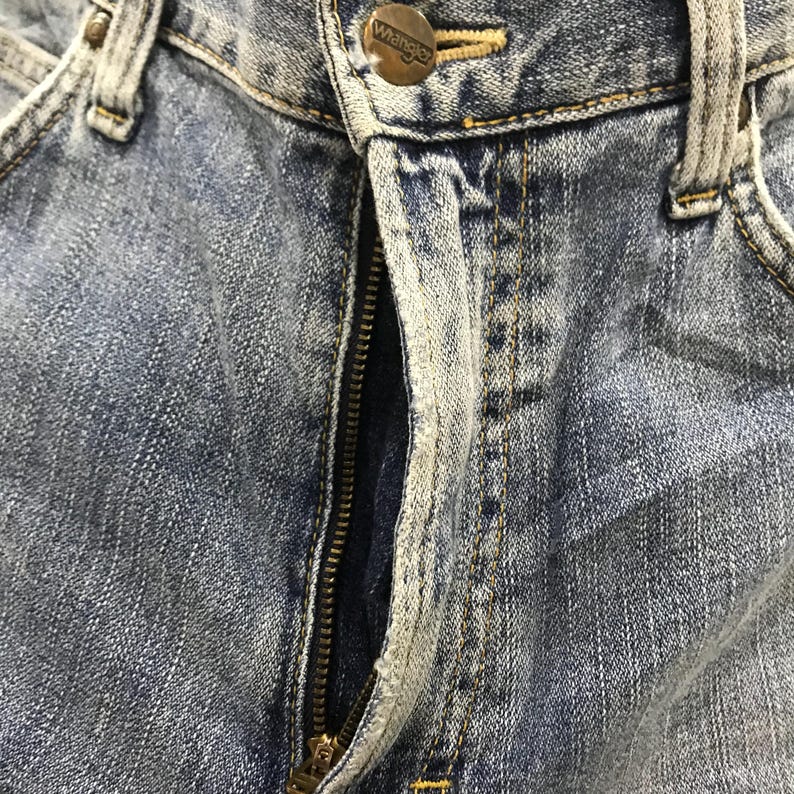 May include: Close-up of blue denim jeans with a visible zipper and a button closure. The jeans show signs of wear and fading, with a metal button that reads "Wrangler".