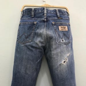 Op de afbeelding: Verbleekte blauwe denim jeans met distressed details, waaronder een grote scheur op de rug. De jeans hebben een klassieke vijf-pocket stijl, een Wrangler merkpatch en een metalen knoopsluiting. De jeans hangen aan een houten hanger.