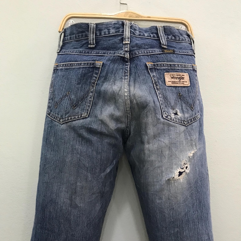 Op de afbeelding: Verbleekte blauwe denim jeans met distressed details, waaronder een grote scheur op de rug. De jeans hebben een klassieke vijf-pocket stijl, een Wrangler merkpatch en een metalen knoopsluiting. De jeans hangen aan een houten hanger.