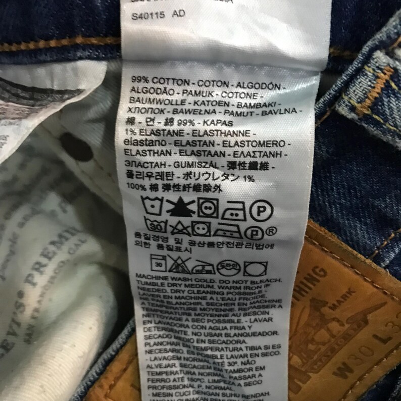 May include: Close-up of a Levi's jeans tag with care instructions and fabric composition details. The tag includes text in multiple languages, detailing washing and ironing guidelines. The jeans are a dark blue denim.