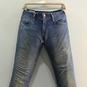 May include: Faded blue denim jeans with a distressed look, featuring a worn appearance and a ripped knee. The jeans are displayed on a wooden hanger against a neutral background. Classic five-pocket design.