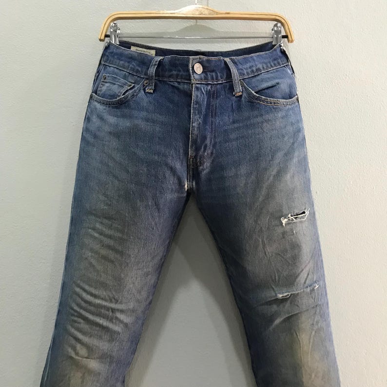 May include: Faded blue denim jeans with a distressed look, featuring a worn appearance and a ripped knee. The jeans are displayed on a wooden hanger against a neutral background. Classic five-pocket design.