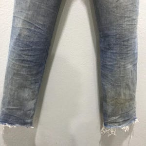 以下が含まれることがあります： 色褪せたダメージ加工が施されたライトブルーのデニムジーンズ。足首には切りっぱなしのフリンジヘム。生地には、明るいパッチやシワなど、目に見える摩耗が見られます。