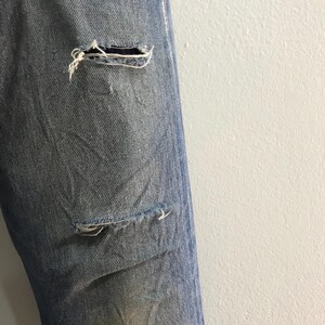 May include: Close-up of light blue denim jeans with distressed details. The jeans have two visible tears, one near the knee and another lower down. Frayed edges and visible threads add to the worn aesthetic of the garment.