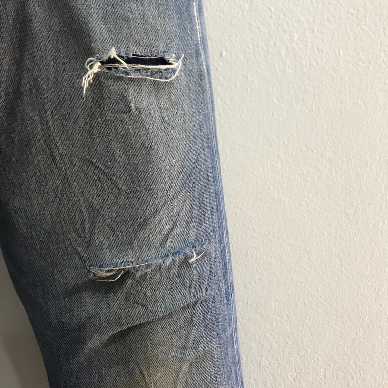 May include: Close-up of light blue denim jeans with distressed details. The jeans have two visible tears, one near the knee and another lower down. Frayed edges and visible threads add to the worn aesthetic of the garment.